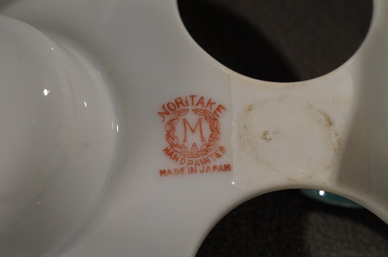 May include: Close-up of a white ceramic egg holder with a red and white Noritake logo that reads "Noritake Hand Painted Made in Japan".