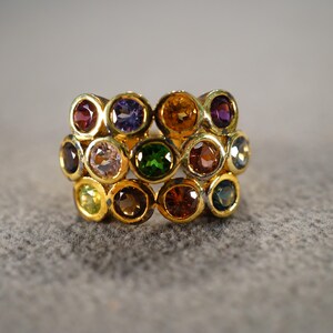 May include: A gold ring with a band of multi-colored gemstones. The stones are set in a circular pattern and include red, orange, yellow, green, blue, and purple.