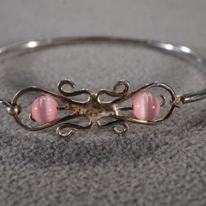 May include: A silver bangle bracelet with a delicate, ornate design featuring two pink cat's eye stones.