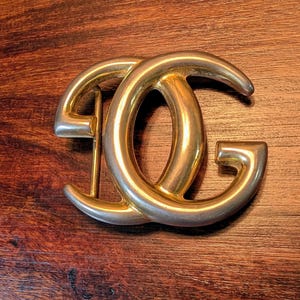 May include: A gold-toned metal belt buckle featuring an interlocking circular design that forms the letters D and G. The buckle has a smooth, polished finish with a curved, rounded frame and a vertical metal bar on the left side for attachment.