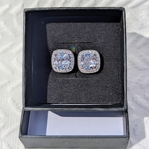 May include: A pair of square-shaped stud earrings featuring a large central clear stone surrounded by a halo of smaller clear stones, set in a silver-toned metal, presented inside a black square jewelry box with a black foam insert.