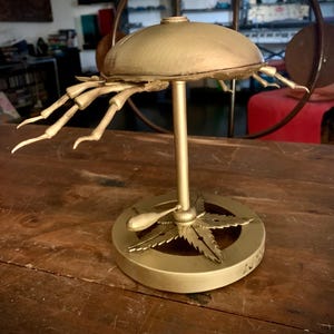 Op de afbeelding: Een gouden metalen sculptuur van een gestileerd marihuanablad met een ronde bovenkant en drie poten. Het beeld staat op een houten oppervlak.