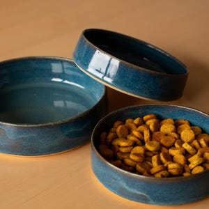 Peut inclure: Trois bols en céramique bleus, dont un rempli de croquettes jaunes pour chien. Les bols sont tous de la même taille et de la même forme, avec un bord légèrement surélevé.