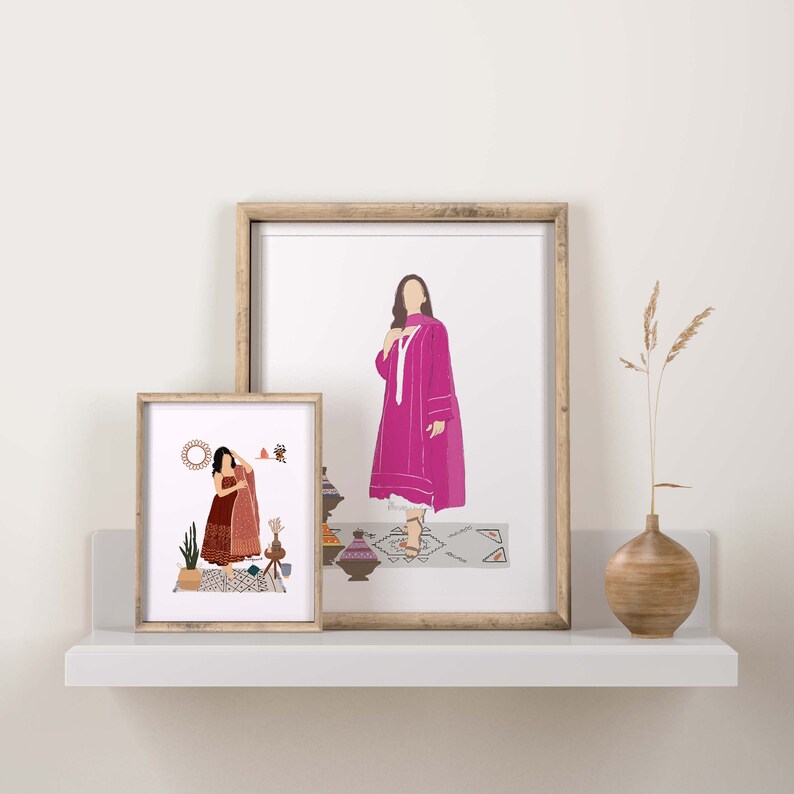 May include: Two framed art prints featuring illustrations of figures in traditional clothing. The larger print shows a woman in a pink outfit, while the smaller one depicts a woman in a red outfit. The prints are displayed on a white shelf with a vase.