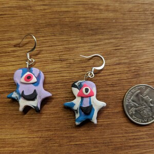 Op de afbeelding: Een paar handgemaakte polymer clay oorbellen met een blauwe, paarse en witte swirl-design met een zwart oog en een roze pupil. De oorbellen zijn bevestigd aan zilveren haakjes.