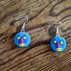 Op de afbeelding: Een paar blauwe oorbellen met een cartoon-gezichtsontwerp. Het gezicht heeft gele ogen, een paarse neus en een gele mond. De oorbellen zijn gemaakt van polymer clay en hebben zilveren haakjes.