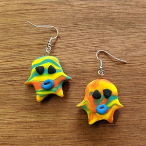Op de afbeelding: Een paar kleurrijke spookvormige oorbellen met blauwe, gele, oranje en groene swirls. De spoken hebben zwarte ogen en een blauwe mond.