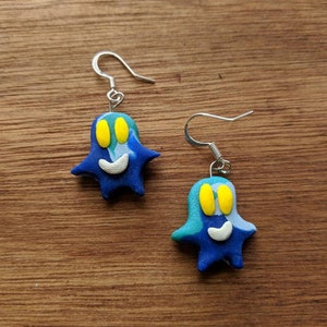 Op de afbeelding: Een paar blauwe en groene spookvormige oorbellen met gele ogen en een witte glimlach. De oorbellen zijn gemaakt van polymer clay en hebben zilveren haakjes.