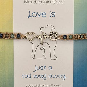 May include: A beige cord bracelet with blue and green speckled beads featuring a silver-toned metal charm of a heart, a heartbeat line, and a paw print. The bracelet is displayed on a white card with the text Island Inspirations, Love is just a tail wag away, and coastalshellcraft.com.
