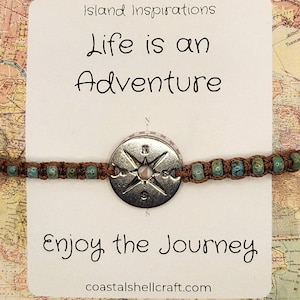 May include: A bracelet with a compass charm and turquoise beads. The card behind the bracelet reads "Life is an Adventure" and "Enjoy the Journey". The background is a map.
