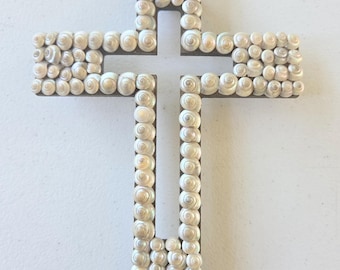 Könnte beinhalten: Ein kreuzförmiges Wandobjekt, das mit kleinen, spiralförmigen weißen Muscheln bedeckt ist. Das Kreuz ist auf einem rechteckigen, hellbraunen Holzsockel montiert. Die Muscheln sind in einem dichten, gleichmäßigen Muster auf der Vorderseite angeordnet.
