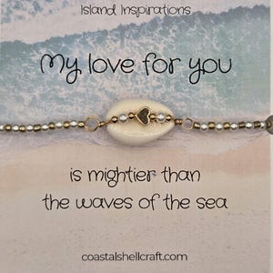 May include: A seashell bracelet with a heart charm, strung with gold and pearl beads. The card behind the bracelet reads "My love for you is mightier than the waves of the sea" with "Island Inspirations" at the top and "coastalshellcraft.com" at the bottom.
