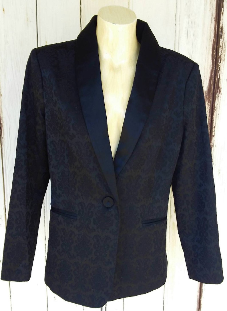 May include: A black blazer with a satin collar and a floral pattern. The blazer has a single button closure.