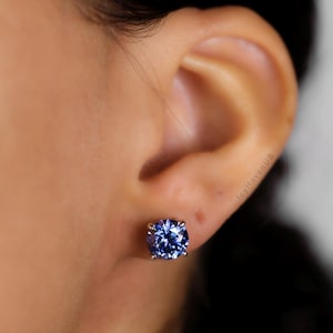 May include: A close-up of a blue gemstone stud earring. The round-cut gemstone is set in a silver or white gold setting. The earring is worn on an ear.
