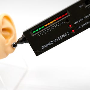 May include: A black diamond tester with a green, yellow, orange, and red LED display. The tester has a small, pointed tip and the words "DIAMOND SELECTOR II" are printed on the side. The tester is being used to test a diamond on a model ear.