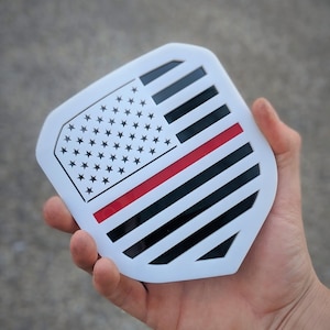 Puede incluir: Una calcomanía blanca en forma de escudo con una línea roja en el medio y un diseño de bandera estadounidense en blanco y negro.