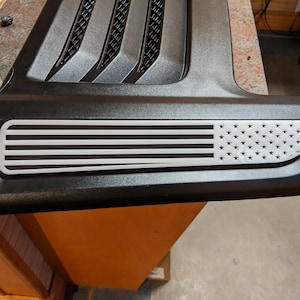 Puede incluir: Una pieza de coche de plástico negro con un diseño de bandera americana blanca. La bandera está en relieve con estrellas y rayas.