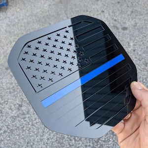 Puede incluir: Emblema de coche de acrílico negro con un recorte de la bandera estadounidense y una fina línea azul.