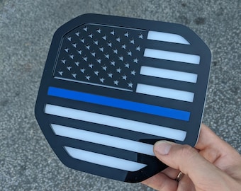 Può includere: Una bandiera americana in bianco e nero con una linea blu che corre orizzontalmente al centro della bandiera. La bandiera è realizzata in un materiale rigido e lucido e ha una forma leggermente arrotondata.