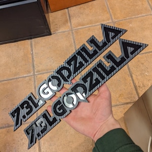 Può includere: Due adesivi "Godzilla" neri e argentati con un motivo in fibra di carbonio. Gli adesivi sono tenuti nella mano di una persona.