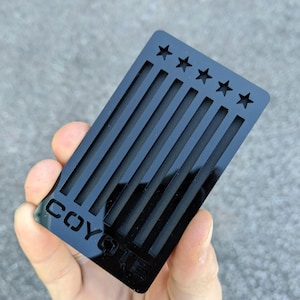Puede incluir: Tarjeta de visita de acrílico negro con un diseño de rayas verticales y estrellas. El texto "COYOTE" está grabado en la tarjeta.