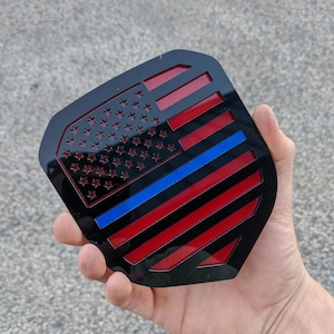 Puede incluir: Emblema negro y rojo con una franja azul, que recuerda a la bandera estadounidense. El emblema tiene un acabado brillante y se sostiene en una mano.