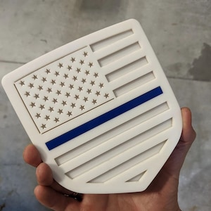 Puede incluir: Placa blanca en forma de escudo con una línea azul y un diseño de bandera estadounidense. La bandera tiene 50 estrellas blancas sobre un campo azul y 13 rayas rojas y blancas.