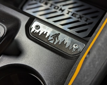 May include: A black and silver Bigfoot-themed car accessory. The metal plate features a silhouette of Bigfoot in a forest scene. The background has the text "CUSTOM TEXT". The accessory is mounted with screws.