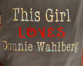 Könnte beinhalten: Graues T-Shirt mit dem Text "This Girl LOVES Donnie Wahlberg" in silbernen und roten Buchstaben. Das Wort "LOVES" hat ein Herzsymbol. Ein lässiges Kleidungsstück.