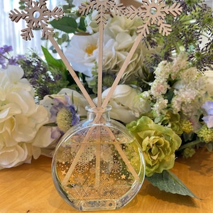 Può includere: Un flacone diffusore rotondo in vetro trasparente con un motivo a fiocco di neve, contenente un liquido chiaro. Bastoncini di legno a forma di fiocco di neve si estendono dal flacone. Lo sfondo presenta un bouquet di fiori bianchi, verdi e viola, con un tema invernale.