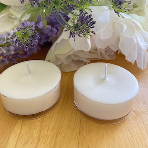 Puede incluir: Dos velas de té blancas con mechas, en soportes de plástico transparente, sobre una superficie de madera. Detrás, flores moradas y blancas crean un fondo suave. Las velas no tienen perfume.