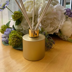 May include: Gold diffuser bottle with a gold lid and wooden diffuser sticks. The bottle is surrounded by white and green flowers.