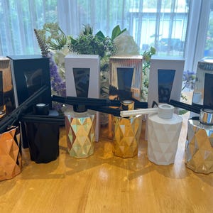 Puede incluir: Ocho botellas difusoras de vidrio decorativas con diseños geométricos en oro, plata y blanco. Las botellas están llenas de aceite perfumado y tienen cañas que sobresalen de ellas.