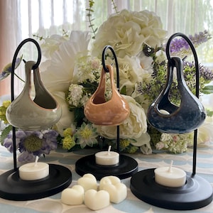 Könnte beinhalten: Drei Keramik-Duftlampen in Grün, Pfirsich und Blau, jeweils an einem schwarzen Metallständer aufgehängt. Weiße Teelichter stehen unter jeder Duftlampe auf einem schwarzen Sockel. Herzförmige Wachsschmelzen sind davor.