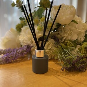 May include: A grey glass diffuser bottle with a silver cap and black diffuser sticks. The bottle is surrounded by a bouquet of white and green flowers.