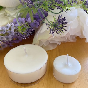 Peut inclure: Deux bougies chauffe-plat blanches avec des m&egrave;ches, l'une plus grande que l'autre, sont pos&eacute;es sur une surface en bois. Les bougies sont dans des supports en m&eacute;tal argent&eacute;. De la lavande violette et des fleurs blanches en arri&egrave;re-plan.