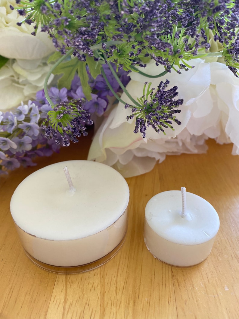Peut inclure: Deux bougies chauffe-plat blanches avec des m&egrave;ches, l'une plus grande que l'autre, sont pos&eacute;es sur une surface en bois. Les bougies sont dans des supports en m&eacute;tal argent&eacute;. De la lavande violette et des fleurs blanches en arri&egrave;re-plan.