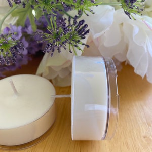 Peut inclure: Deux bougies chauffe-plat blanches avec m&egrave;ches, dans des coupelles en plastique transparent, pos&eacute;es sur une surface en bois. Des fleurs violettes et blanches sont en arri&egrave;re-plan. Les bougies sont non parfum&eacute;es.