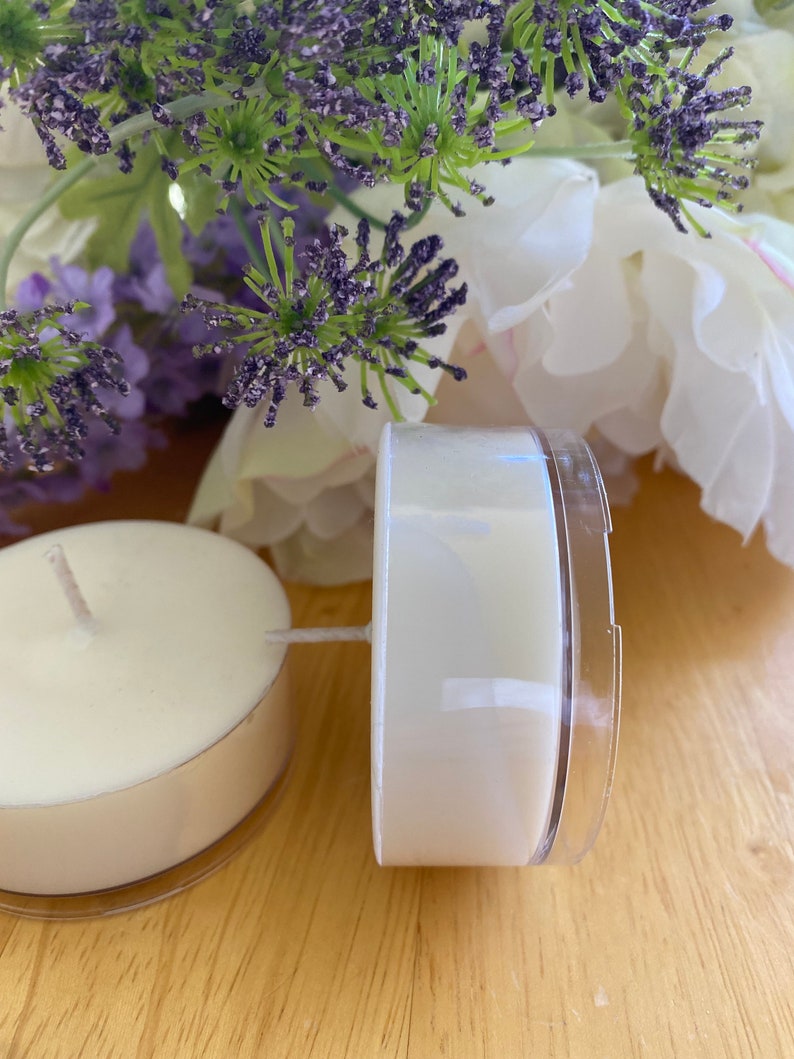 Peut inclure: Deux bougies chauffe-plat blanches avec m&egrave;ches, dans des coupelles en plastique transparent, pos&eacute;es sur une surface en bois. Des fleurs violettes et blanches sont en arri&egrave;re-plan. Les bougies sont non parfum&eacute;es.