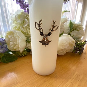 Könnte beinhalten: Eine große, elfenbeinfarbene Säulenkerze mit einem Hirschkopf-Emblem in Bronzetönen. Die Kerze ist von weißen und lila Blumen umgeben. Es ist ein dekorativer Artikel.
