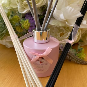 Puede incluir: Una botella de difusor de vidrio rosa con una tapa plateada y un lazo blanco. La botella tiene una etiqueta que dice "Wick Candles Angel www.etsy.com/shop/FlorisHouse". La botella está rodeada de flores blancas y moradas y tiene varios bastones difusores de madera.