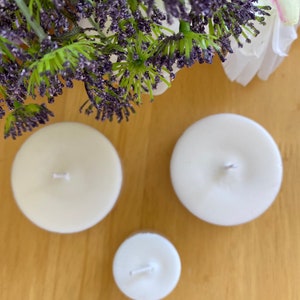 Peut inclure: Trois bougies rondes, non parfum&eacute;es, de couleur ivoire, de tailles diff&eacute;rentes. Chaque bougie a une seule m&egrave;che. Les bougies sont dispos&eacute;es sur une surface en bois brun clair, avec des accents floraux violets et verts en arri&egrave;re-plan.