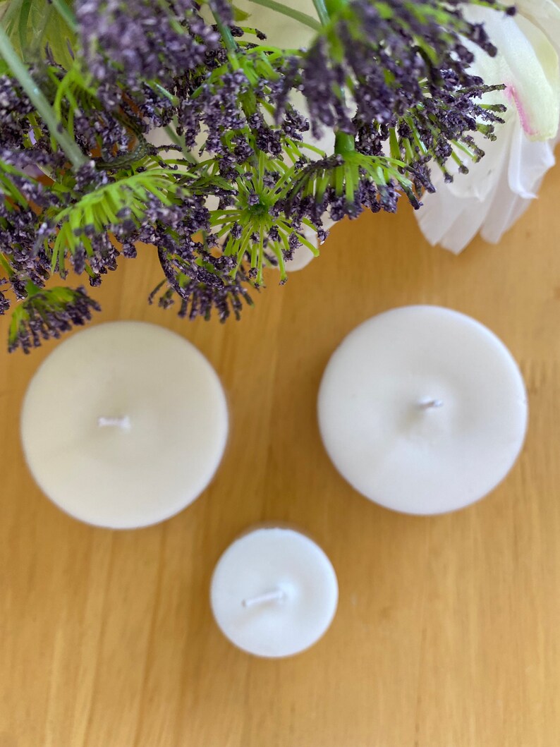 Peut inclure: Trois bougies rondes, non parfum&eacute;es, de couleur ivoire, de tailles diff&eacute;rentes. Chaque bougie a une seule m&egrave;che. Les bougies sont dispos&eacute;es sur une surface en bois brun clair, avec des accents floraux violets et verts en arri&egrave;re-plan.