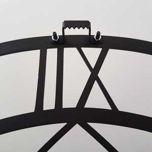 A close-up back view of a matte black Metal Wall Clock, showcasing the sturdy and reliable sawtooth hanger mechanism for easy installation. The durable metal mounting bracket is securely fastened with screws and nuts directly above the precision laser-cut Roman numeral XII. This detailed shot highlights the practical demonstrating premium craftsmanship and safe wall mounting design. Essential product detail for Etsy shoppers seeking high-quality, easy-to-install Viking Decor.