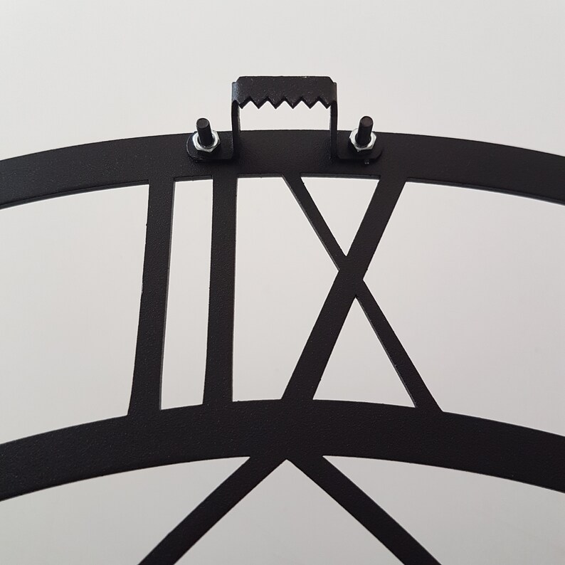 A close-up back view of a matte black Metal Wall Clock, showcasing the sturdy and reliable sawtooth hanger mechanism for easy installation. The durable metal mounting bracket is securely fastened with screws and nuts directly above the precision laser-cut Roman numeral XII. This detailed shot highlights the practical demonstrating premium craftsmanship and safe wall mounting design. Essential product detail for Etsy shoppers seeking high-quality, easy-to-install Viking Decor.