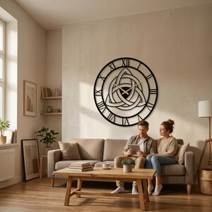 "A curated modern living room featuring a striking large black metal wall clock with intricate Triquetra and Trinity Knot design center and detailed Roman numerals.This Modern Wall Art piece which hangs above a couple relaxing on a sofa, looking at a tablet. Perfect as unique Viking Decor and Home Decoration, suitable for unique gifts and curated decor.