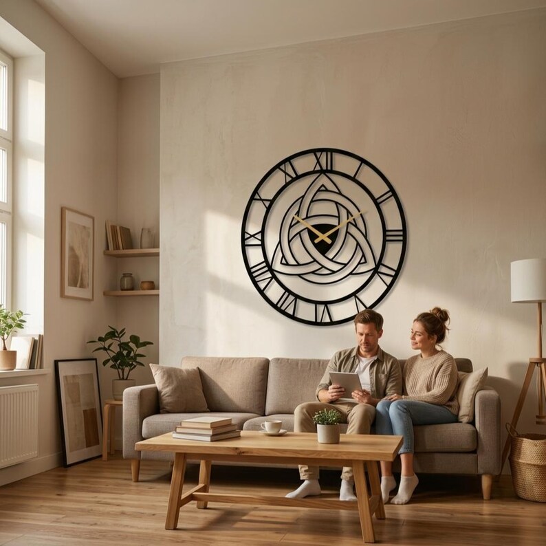 "A curated modern living room featuring a striking large black metal wall clock with intricate Triquetra and Trinity Knot design center and detailed Roman numerals.This Modern Wall Art piece which hangs above a couple relaxing on a sofa, looking at a tablet. Perfect as unique Viking Decor and Home Decoration, suitable for unique gifts and curated decor.