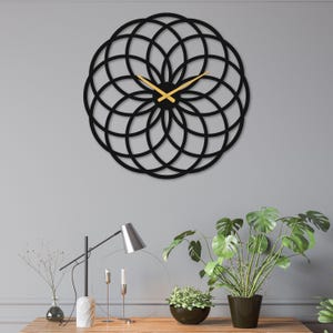 May include: A black, geometric wall clock with a circular design. The clock features gold-colored hands and a modern, minimalist aesthetic. The clock is mounted on a gray wall, above a wooden table with plants and a lamp.