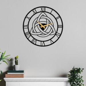 Metal Wall Clock by Degary, featuring an intricate central Triquetra Trinity Knot design and elegant Roman numerals with modern gold hands. The clock is mounted on a minimalist light grey wall directly above a white wooden mantelpiece. The clean shelf setup is styled with a small potted cactus, a stack of books, and green indoor plants on either side, showcasing a perfect example of minimalist Nordic Decoration. This striking timepiece serves as authentic Viking Decor and Modern Wall Art.