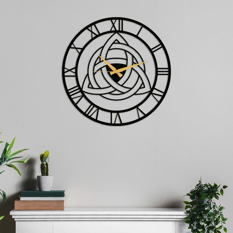Metal Wall Clock by Degary, featuring an intricate central Triquetra Trinity Knot design and elegant Roman numerals with modern gold hands. The clock is mounted on a minimalist light grey wall directly above a white wooden mantelpiece. The clean shelf setup is styled with a small potted cactus, a stack of books, and green indoor plants on either side, showcasing a perfect example of minimalist Nordic Decoration. This striking timepiece serves as authentic Viking Decor and Modern Wall Art.